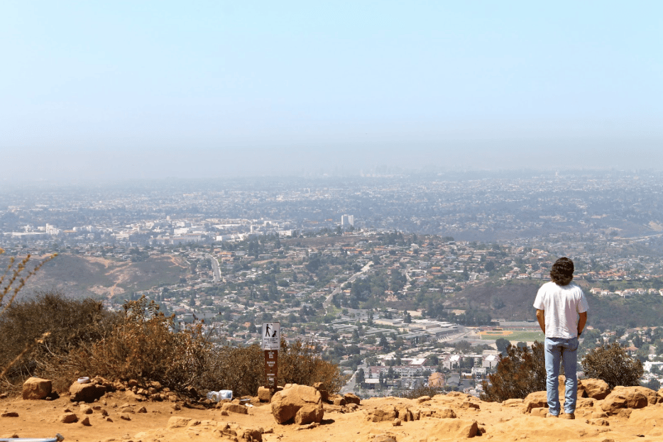 A man wearing a white t-shirt and jeans stands on a rocky hill overlooking San Diego.