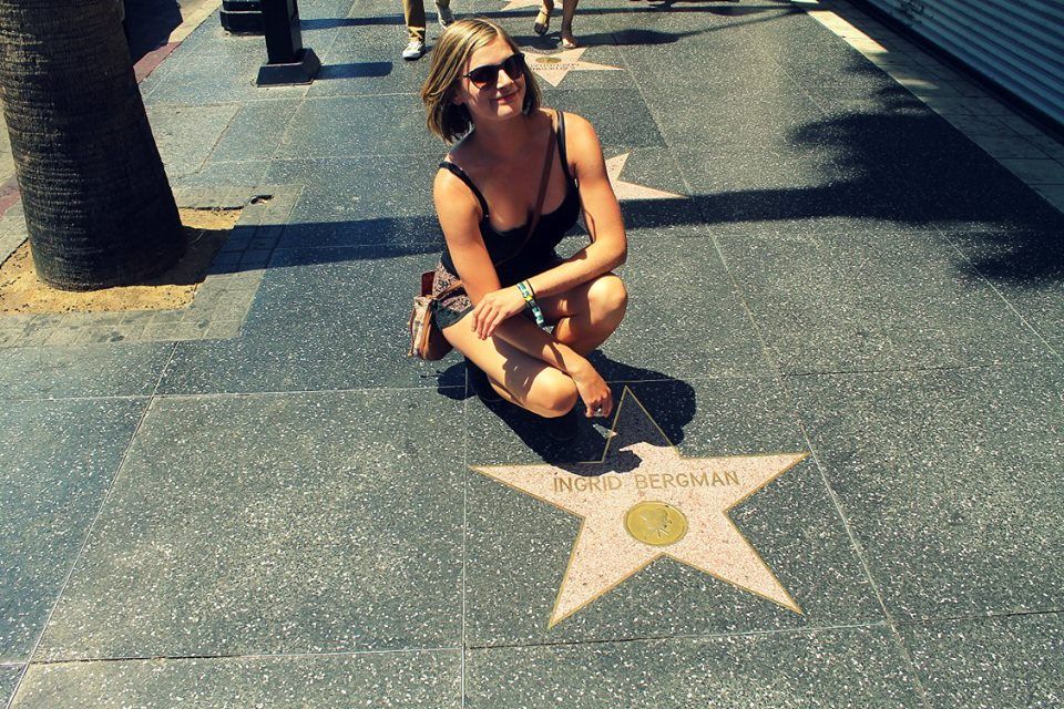 A young woman with short hair and sunglasses crouches, smiling, next to the Ingrid Bergman star on the Hollywood Walk of Fame.