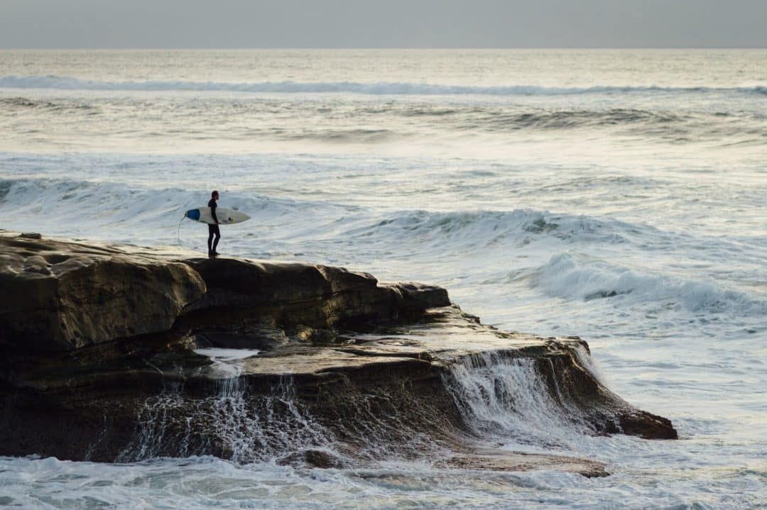 A surfer stands on a rock that sticks out into the sea, surrounded by crashing waves.