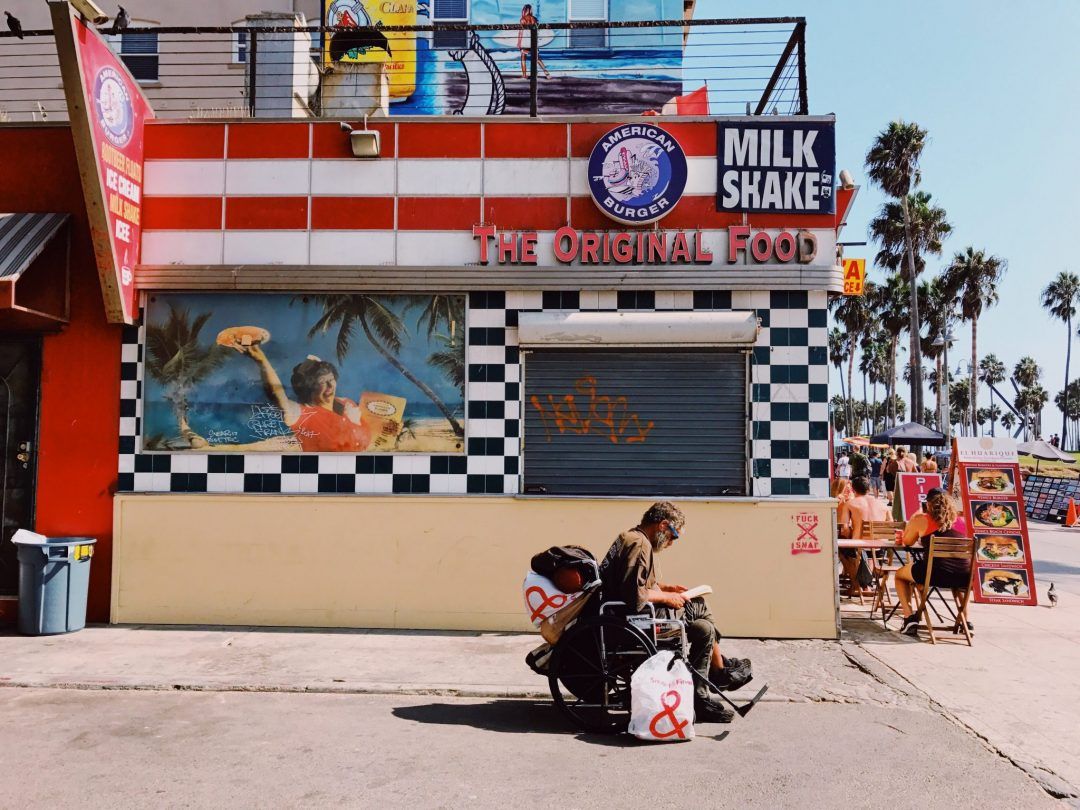 A red, white, and black checkered food stand called American Burger on a beachy street corner in Venice, CA, with palm trees in the background.