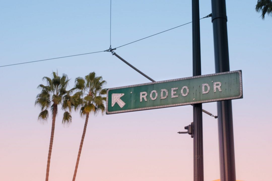 The green street sign of Rodeo Drive against a pink and blue sky at sunset with two palm trees in the background.