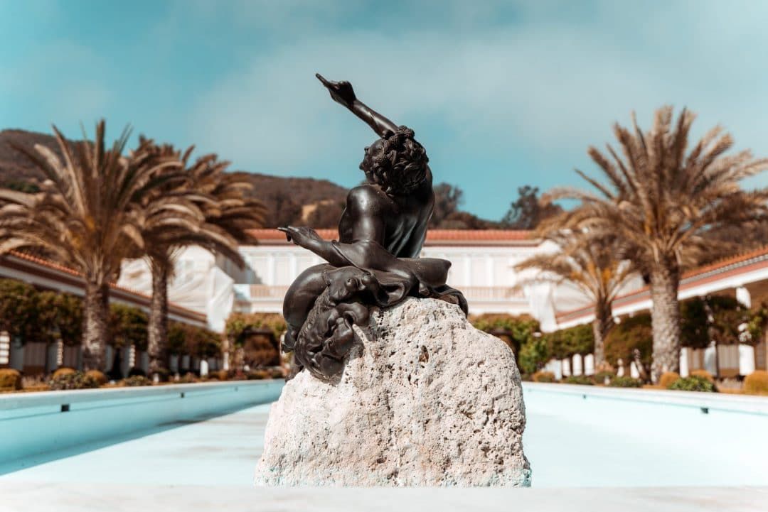 A bronze sculpture of a figure pointing upwards while lounging on a rock with palm trees and blue skies in the background at the Getty Villa.