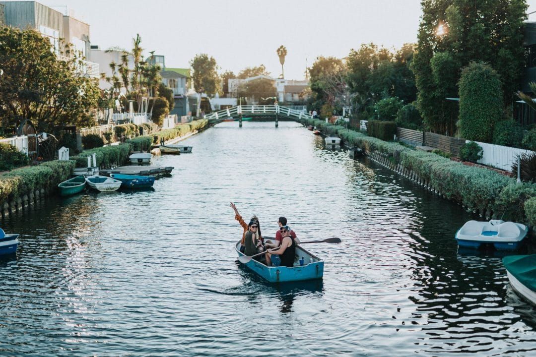 A couple of young people paddling a blue boat down a garden-lined canal in Venice, California.