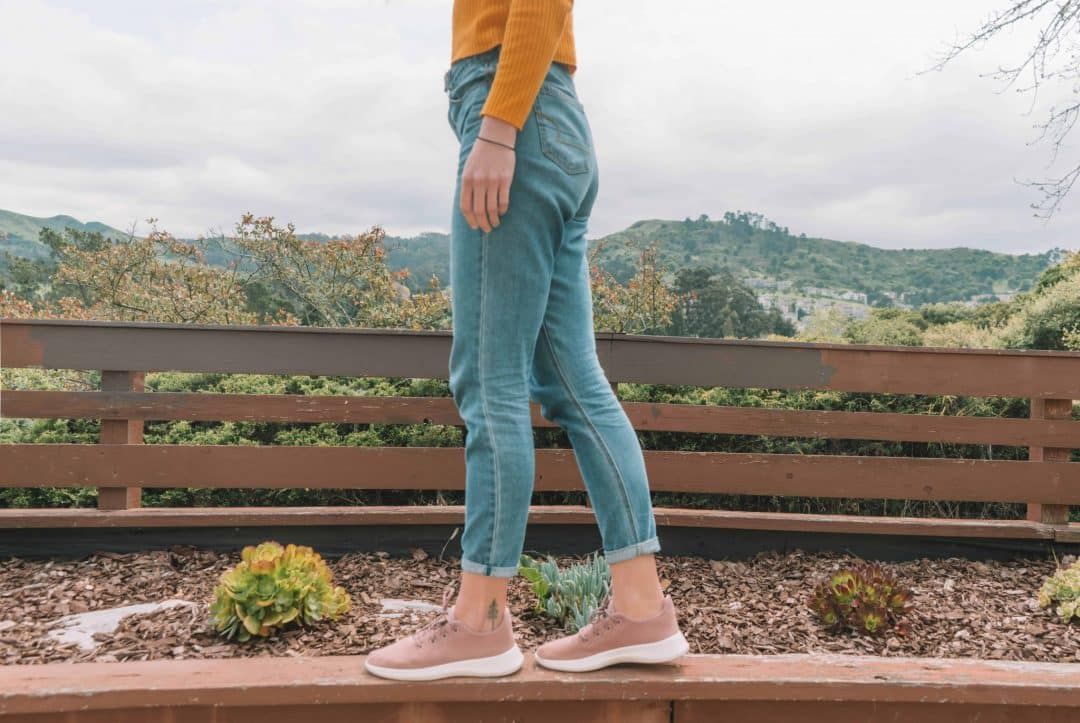 A pair of legs wearing jeans and Allbirds tree runners in pink standing on the edge of a wood planter in a backyard.