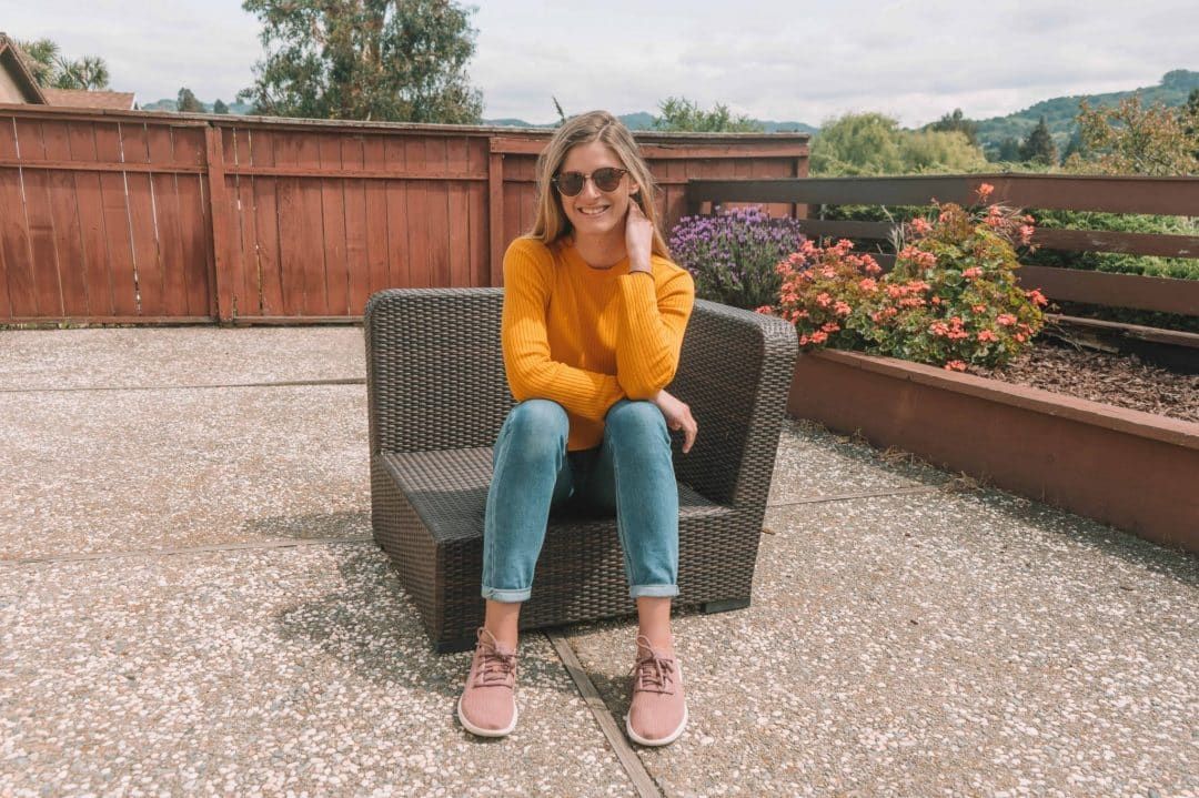 A young woman in jeans, a yellow sweater, sunglasses, and pink Allbirds tree runners smiles and sits on a wicker patio chair.