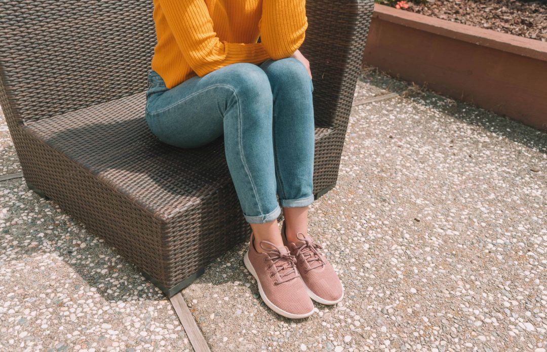 A young woman in jeans, a yellow sweater, and pink Allbirds tree runners seen from the neck down sitting on a wicker patio chair.