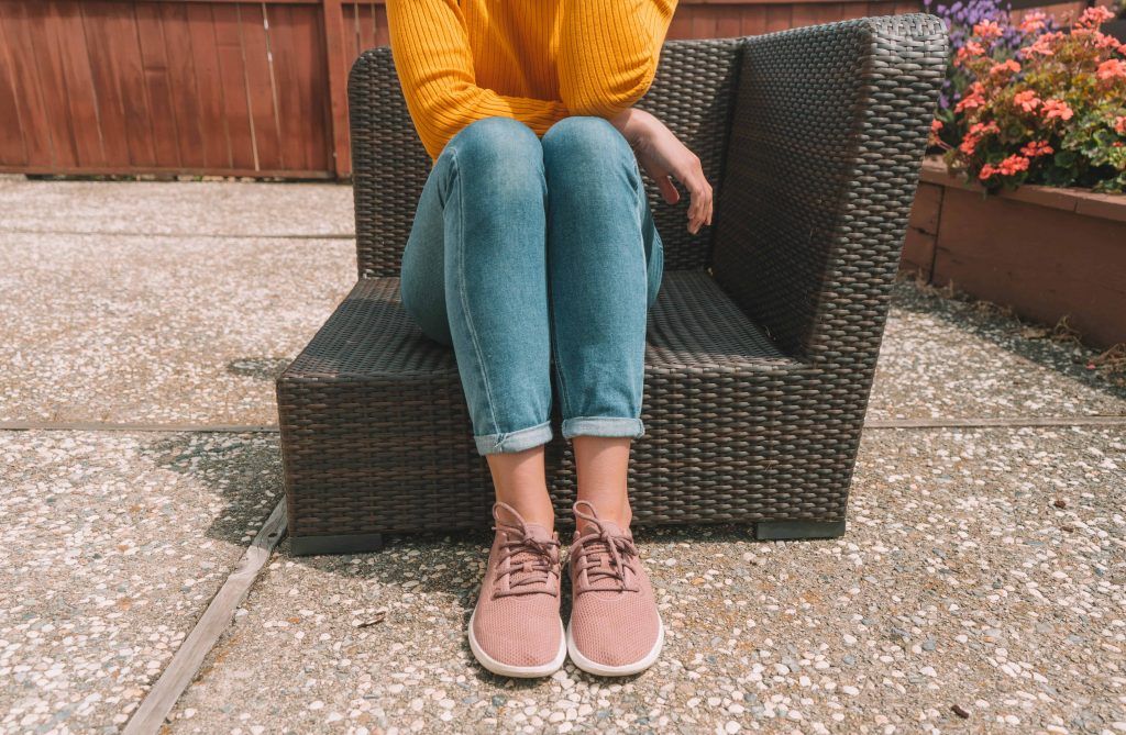 The best Allbirds alternatives: a young woman seen from the neck down wearing a yellow sweater, jeans, and pink Allbirds sitting on a wicker patio lounger with a flowerbed in the background.