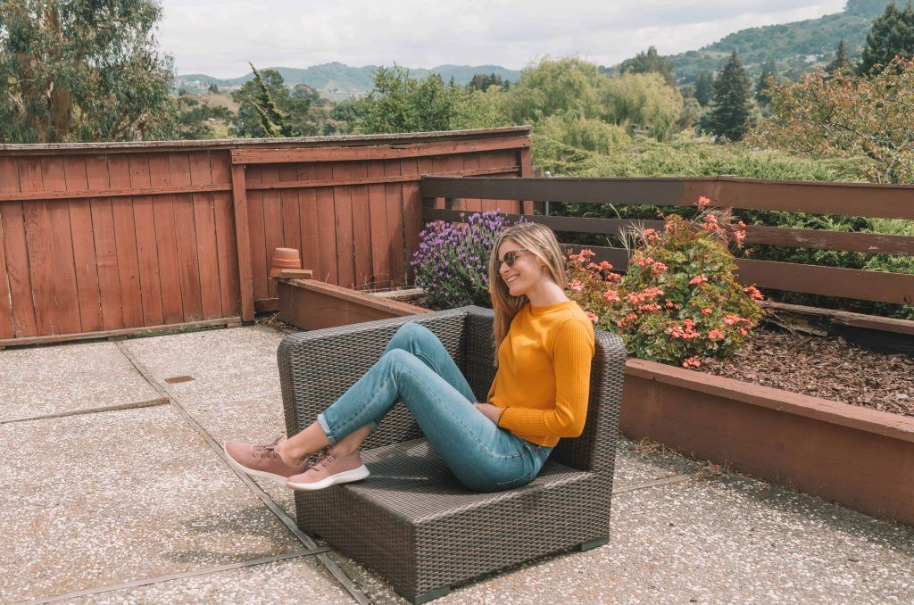 A young woman smiling and wearing sunglasses, a yellow sweater, jeans, and pink Allbirds sitting on a wicker patio lounger with a flowerbed and hills in the background.