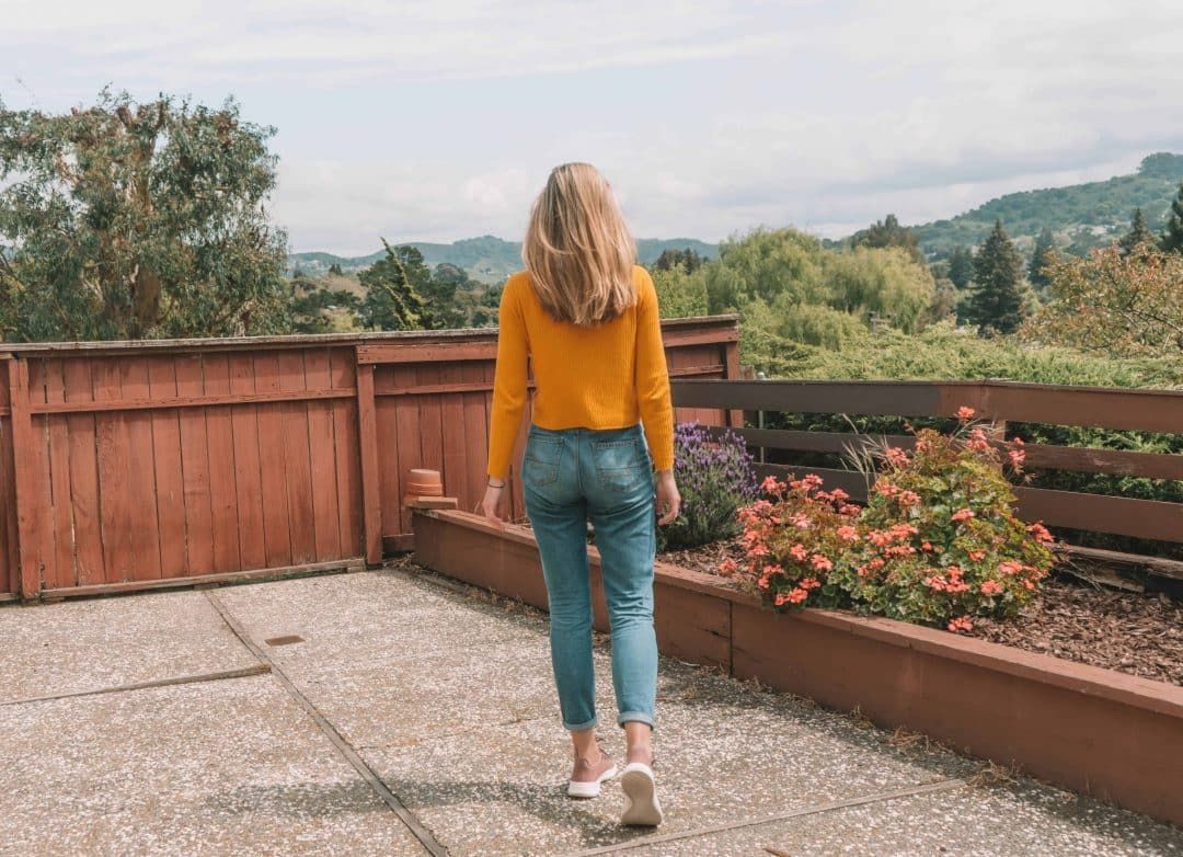 A young woman in jeans, a yellow sweater, and  pink Allbirds tree runners seen from behind on a concrete patio with trees and hills in the background.