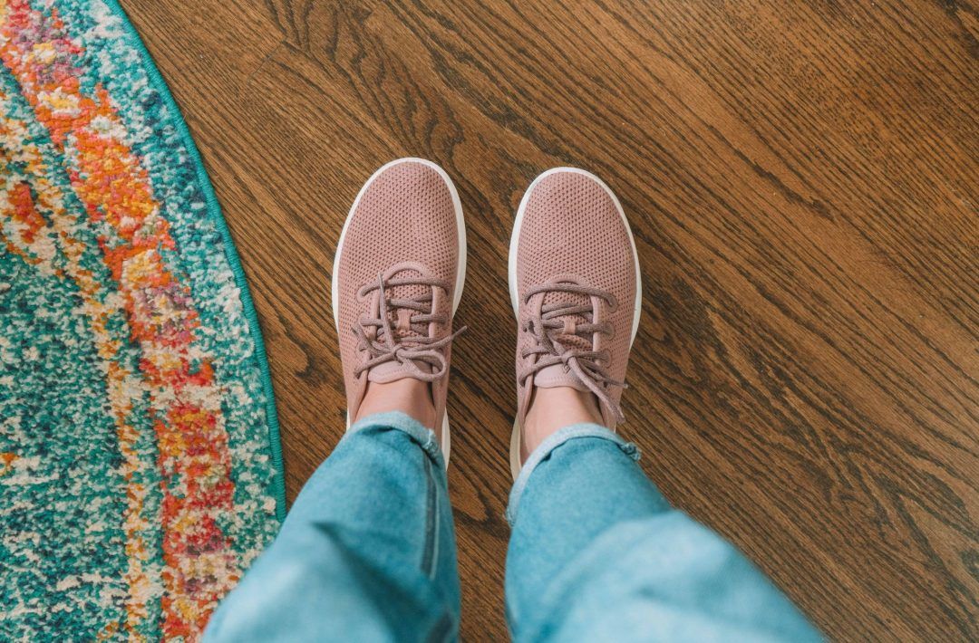 A pair of feet wearing Allbirds tree runners in pink, standing on a hardwood floor next to a colorful rug.