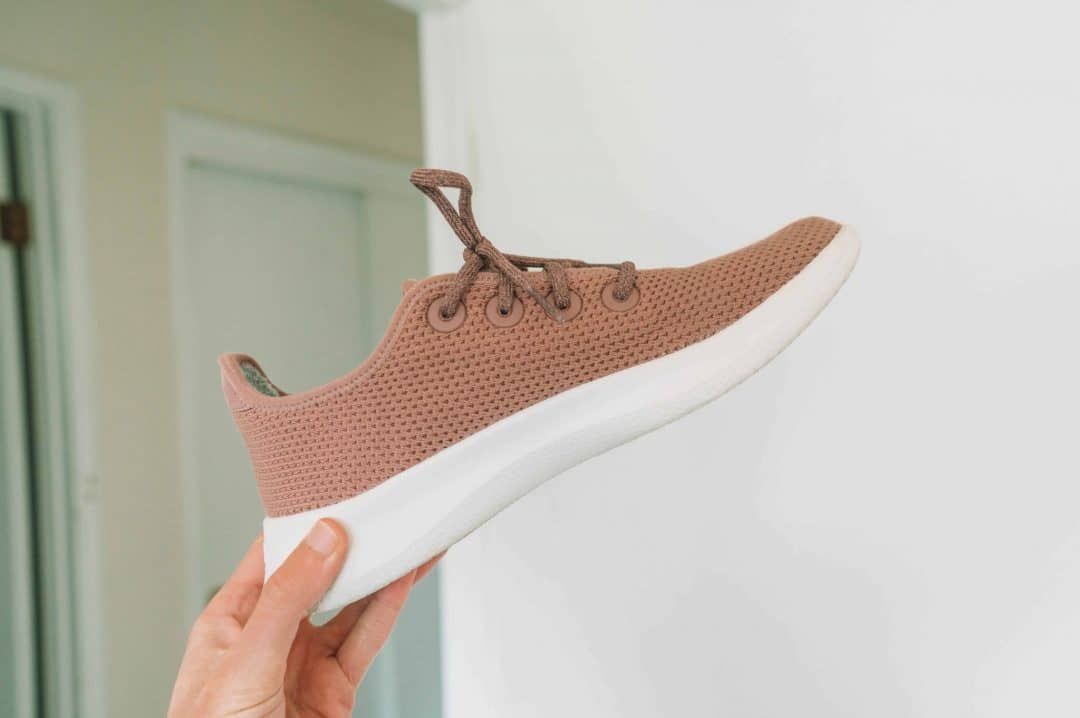 A hand holding up a pink Allbirds tree runners sneaker with a white walled-room in soft focus behind it.