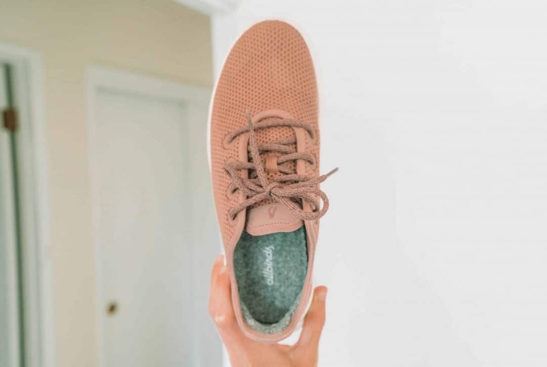 A hand holding up a pink Allbirds tree runners sneaker with a white walled-room in soft focus behind it.
