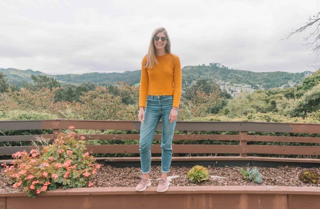 Allbirds shoes review: A young woman in jeans, a yellow sweater, and  pink Allbirds tree runners stands on the edge of a brown planter box with trees and hills in the background.