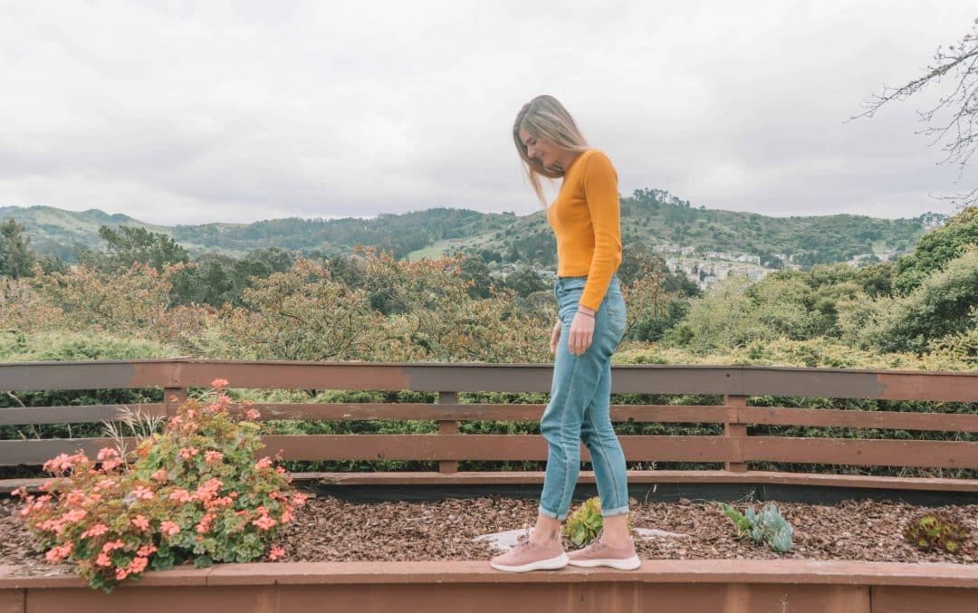 A young woman in jeans, a yellow sweater, and  pink Allbirds tree runners stands on the edge of a brown planter box with trees and hills in the background.