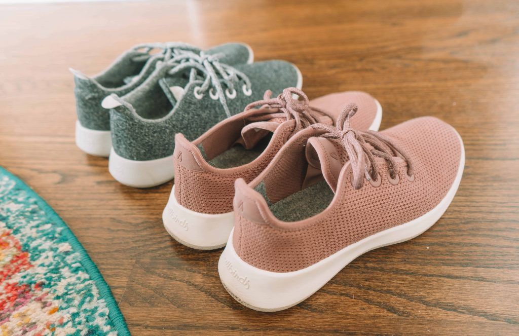 Two pairs of Allbirds shoes, one grey and the other pink, sitting on a hardwood floor next to a colorful circular rug.
