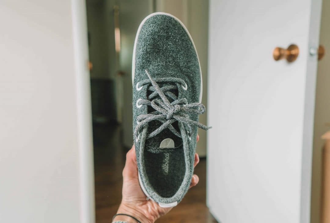 A hand holding up a grey Allbirds wool runner with a white-walled room in soft focus in the background.