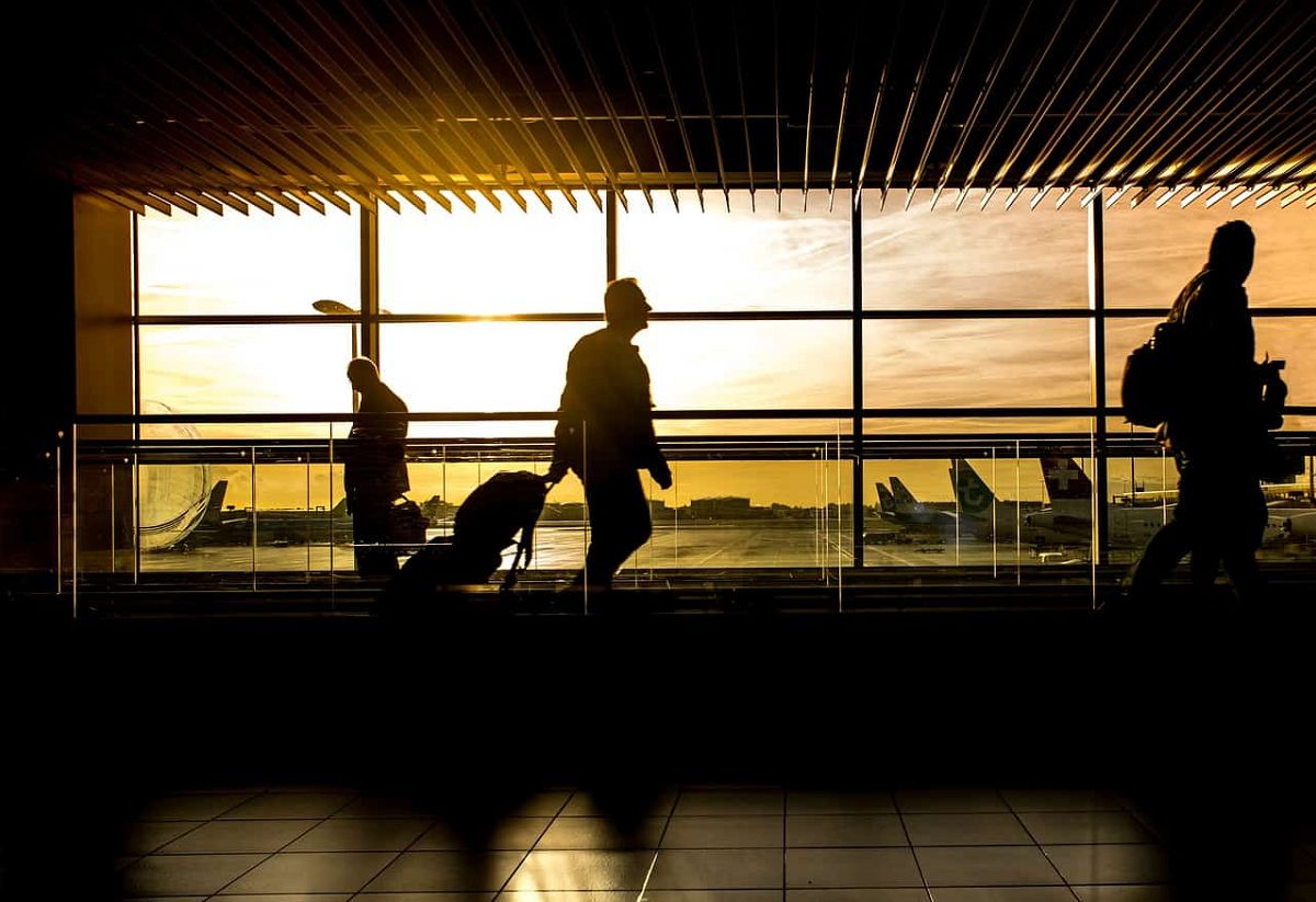 Silhouettes of people on a moving walkway in an airport, with the tarmac and a yellow-orange sunset visible through the window behind them.