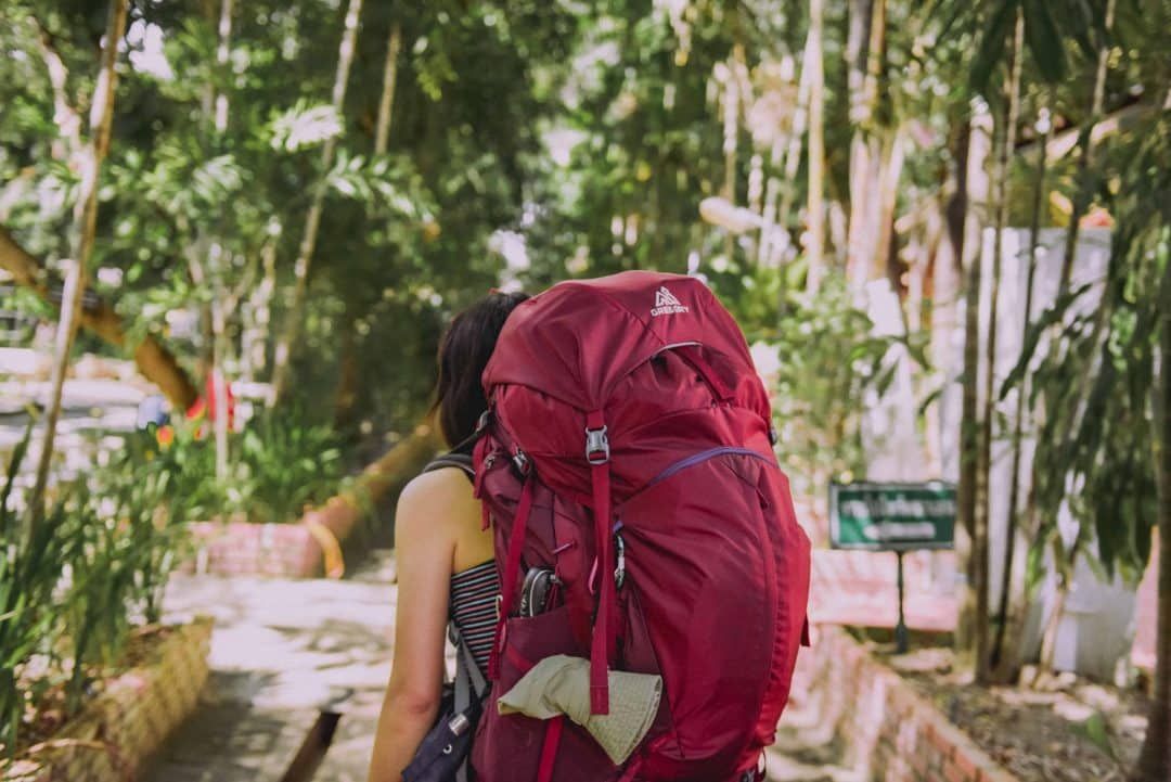 A young, dark-haired woman in a striped shirt seen from behind wearing a large red backpacking backpack in a soft-focus tropical setting.