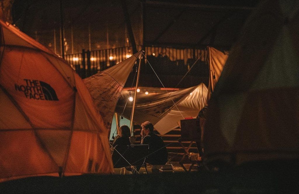 Two people sit in camping chairs amongst tents in a warmly-lit campsite at night.