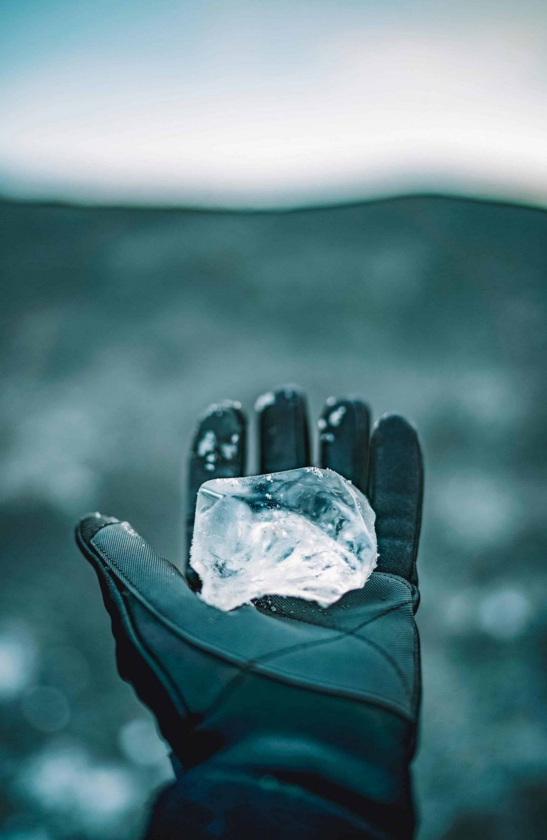 A gloved hand holds out a chunk of ice in front of of a blurred landscape.