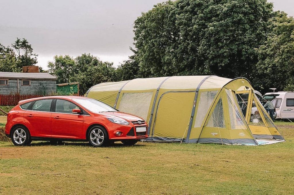 A large, green tunnel-style tent pitched on a grassy lawn beside a red hatchback car, with trees, RVs, and a cabin in the background.