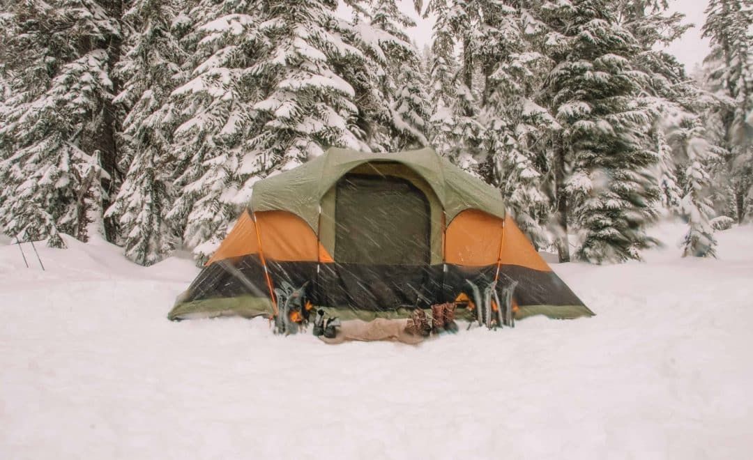 A green, orange, and black tent pitched into the snow during a blizzard with snowy evergreen trees behind it.