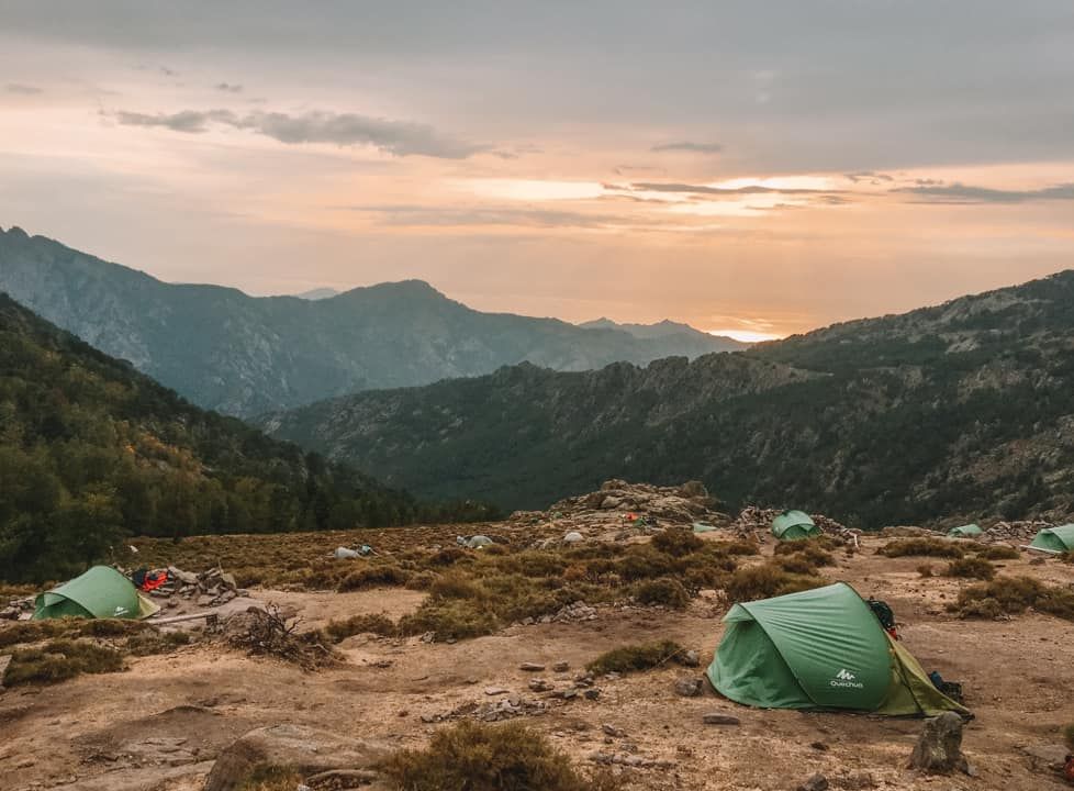 A sparse, mountainous landscape at dusk with a dark green pop-up tent pitched in the forground, with more green tents in the background.