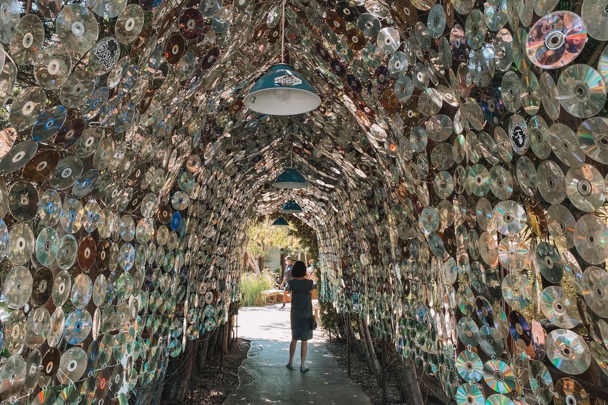 A woman with short dark hair walks through a an art installation consisting of a tunnel lined with hundred of CDs.
