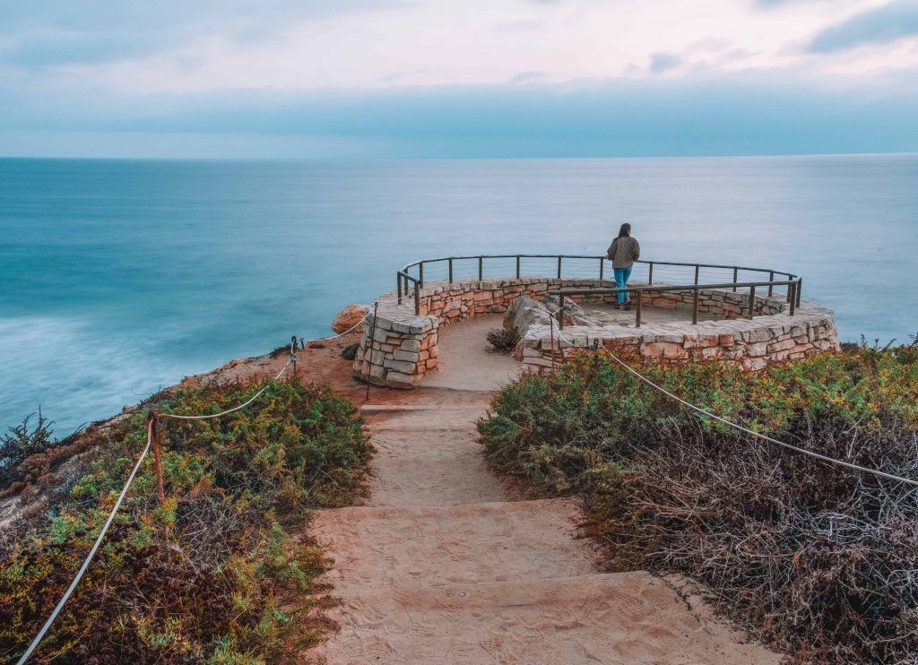 A sandy path leading to a circular stone lookout point where a woman stands at the railing and looks out over the ocean, with an overcast sky above.