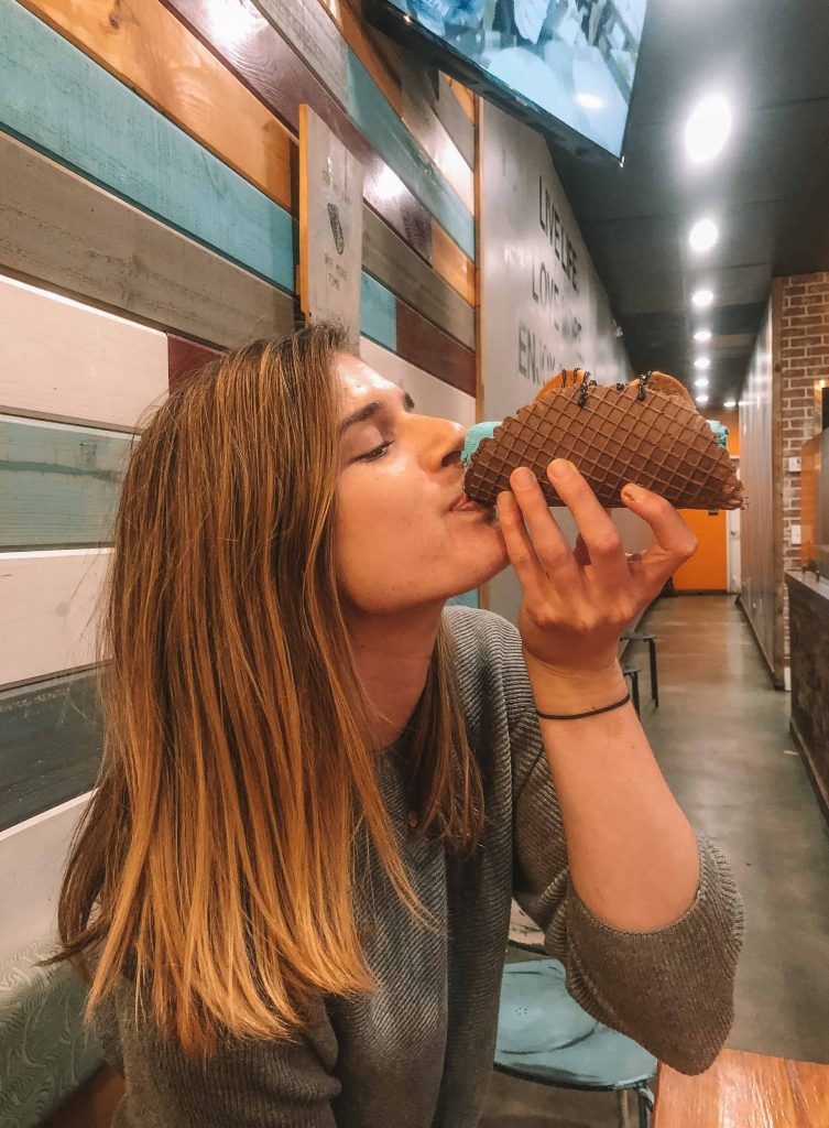 A woman with light brown hair turns to the side to bite into an ice cream taco, with the interior of a brightly-lit ice cream shop in the background.