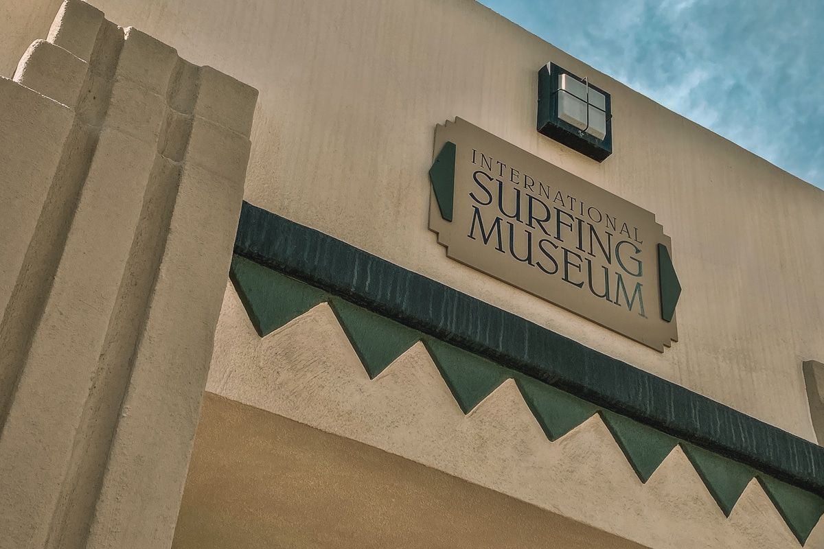 A close-up of the sign for the International Surfing Museum, with some of the building's art-deco facade and a sliver of blue sky visible beyond.