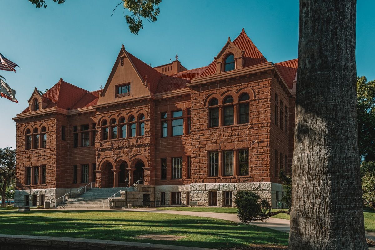 The Old Orange County Courthouse, a large, reddish-brown stone building surrounded by lawn with a tree trunk in the foreground and a blue sky in the background.