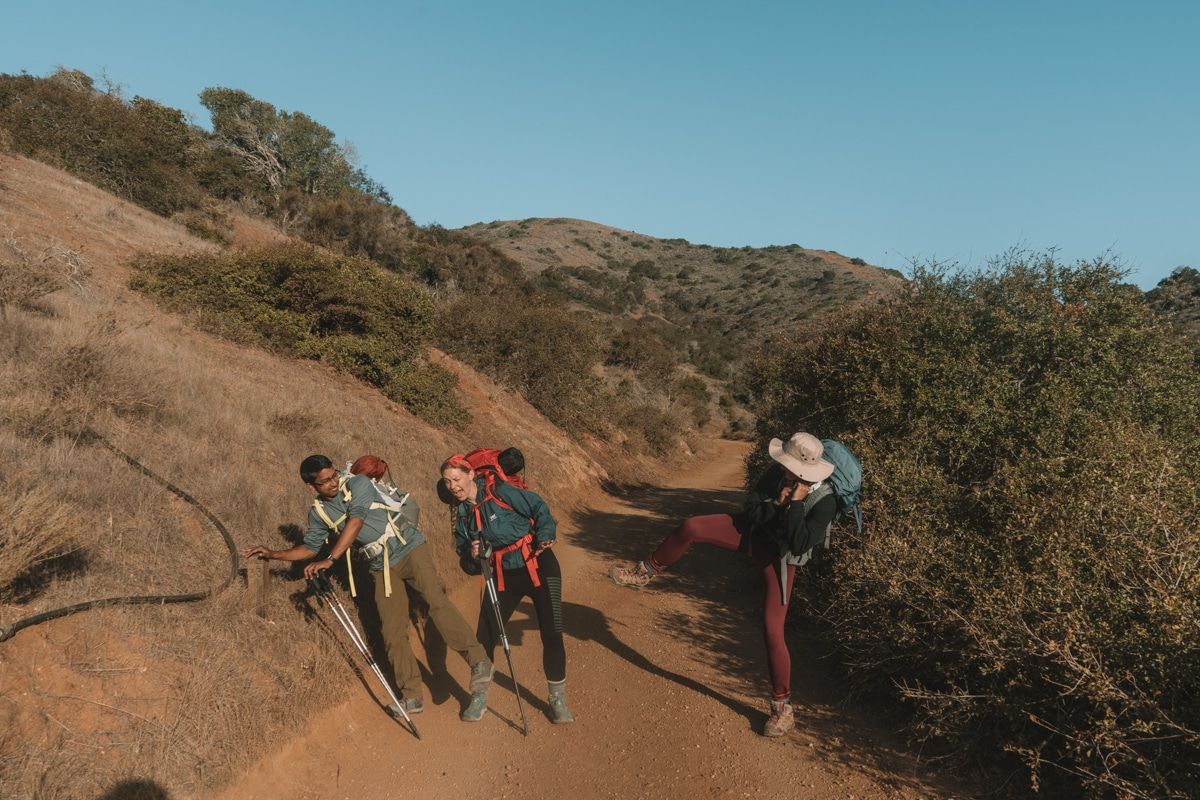 Three friends in hiking gear goofing around on a trail during a backpacking trip across Catalina Island, with California hills and a bright blue sky in the background.