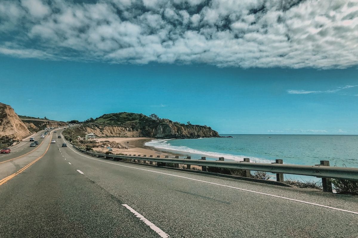 A view of the open road of the Pacific Coast Highway, with a beach to the left and partly cloudy sky above.