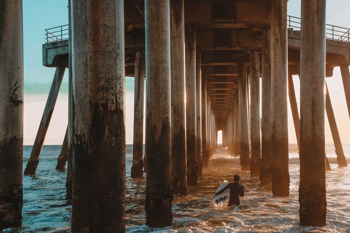 A surfer seen from behind making his way out into the waves underneath a pier, amongst the posts.