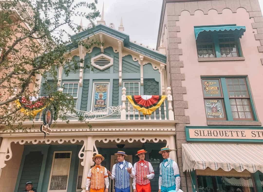 A barbershop quartet dressed in bright colors stands in front of  a Victorian-style wooden facade at Disneyland.