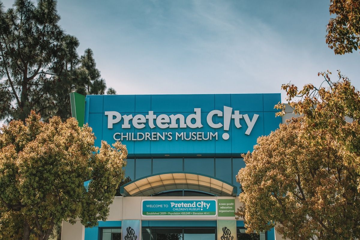 The blue front entrance and sign for the Pretend City Children’s Museum surrounded by trees with a partly cloudy sky in the background.