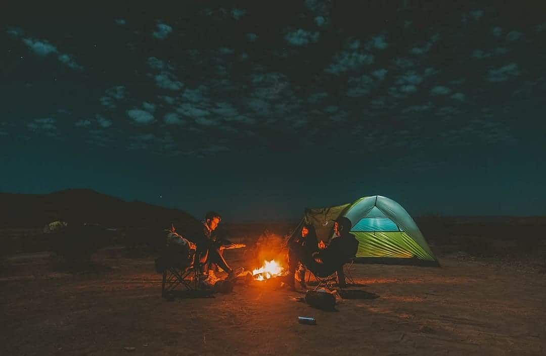 Three people sit around a campfire in a dark, desert landscape, with a blue tent illuminated in the background.