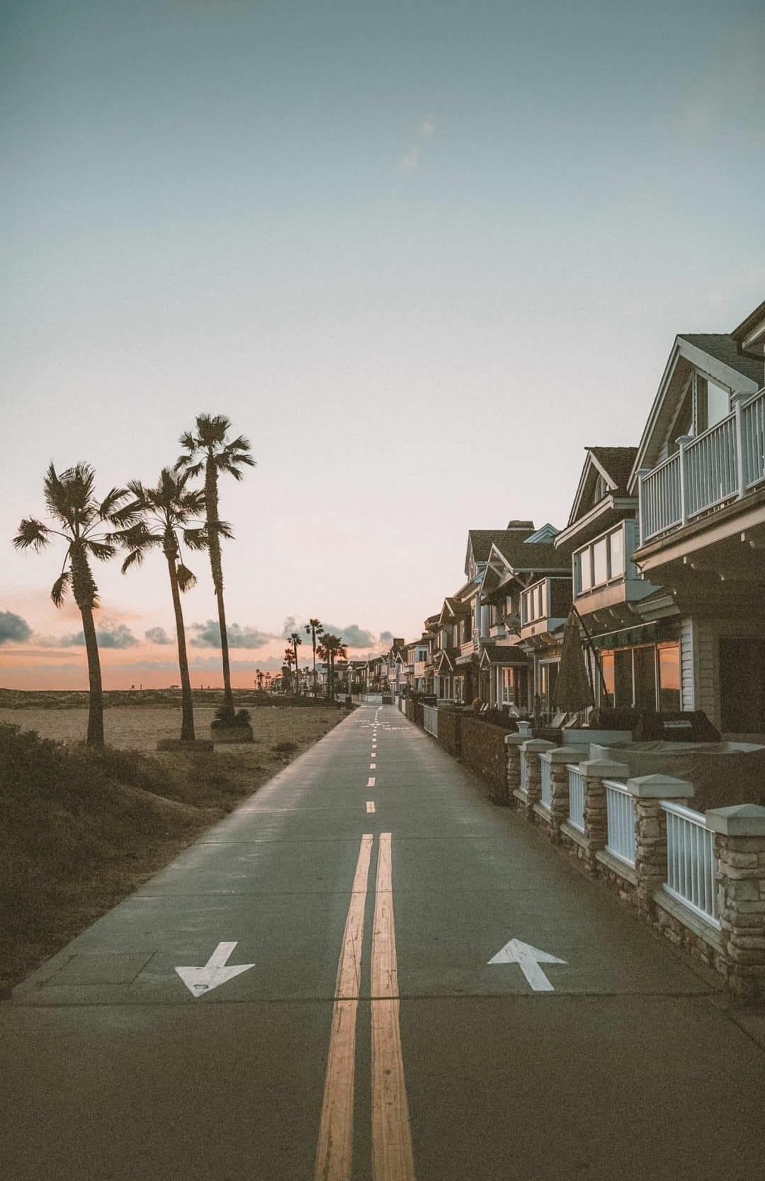 A view of the Newport Beach Bike path at sunset, with three palm trees on the beach to the left, and a row of beach houses to the right.
