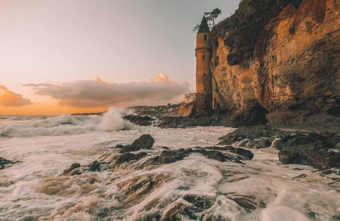 A fairytale-style castle tower built against a sandstone cliff overlooking crashing waves, with a sunset in the distance.