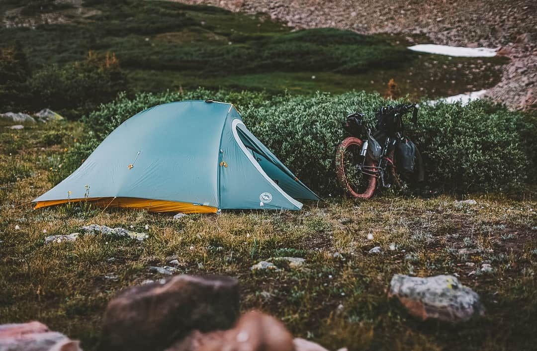 A small, turquoise tent pitched in a damp-looking, rocky landscape, with a mountain bike leaning against a nearby hedge.