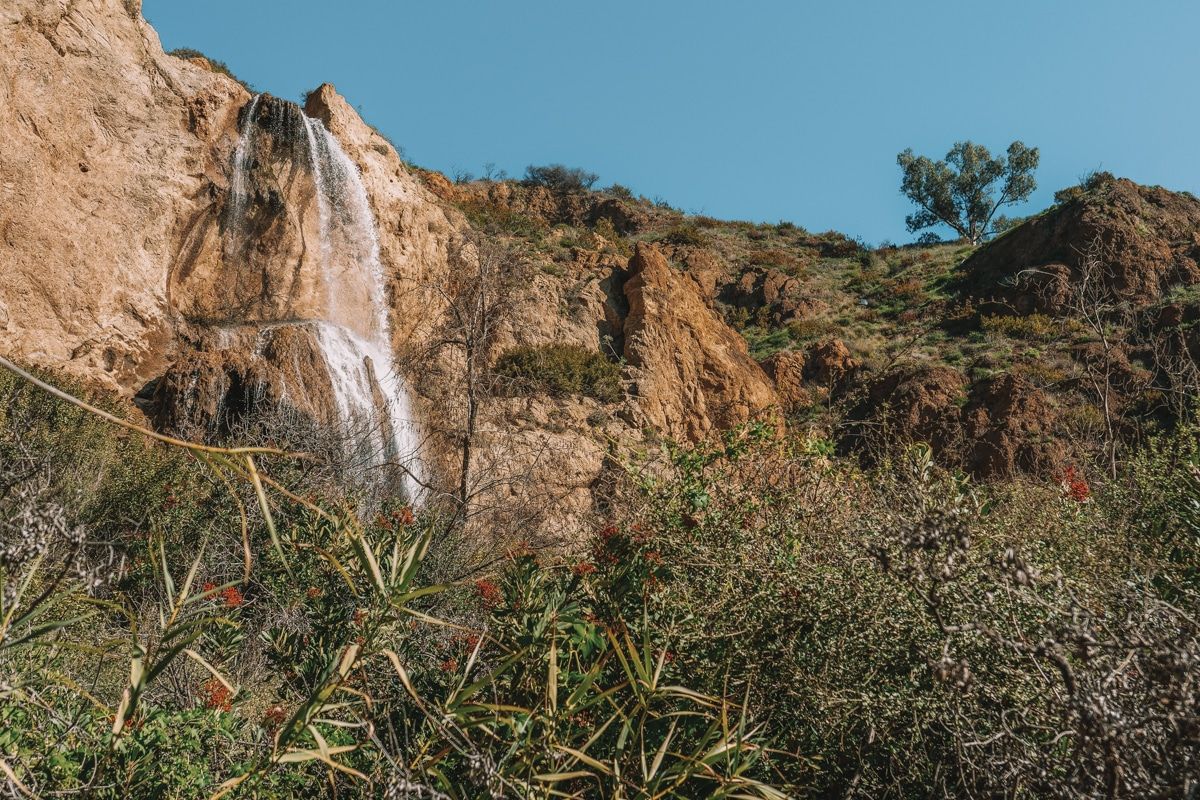 A view from the bushes below of Escondido Falls, a waterfall spilling over reddish rock.