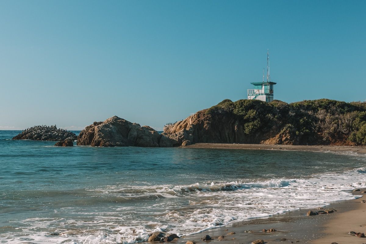 The calm waves at Leo Carrillo State Park, with a small lookout house visible on top of a rocky dune, with a clear blue sky beyond.