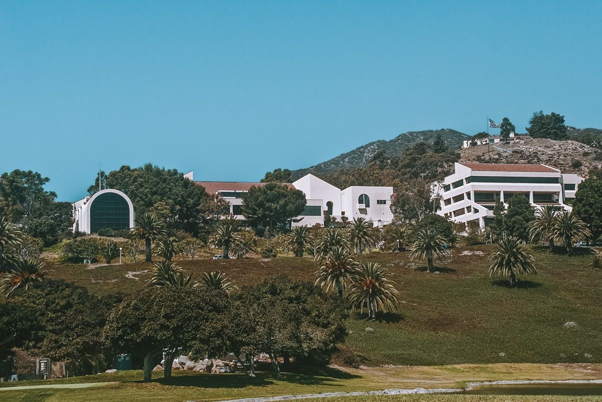 The white buildings of the Pepperdine University campus stand out against green lawns and palm trees, with a bright blue sky beyond.