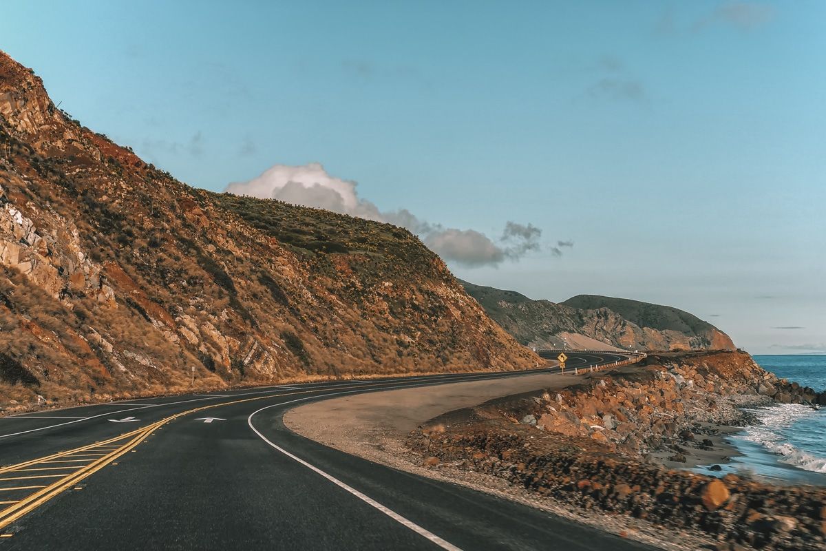 A view of the open road of the Pacific Coast Highway alongside the beach on a clear day.