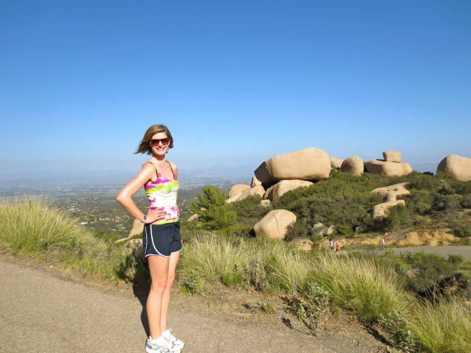 A woman wearing shorts and sunglasses poses in front of some canyon rock formations against a clear blue sky.