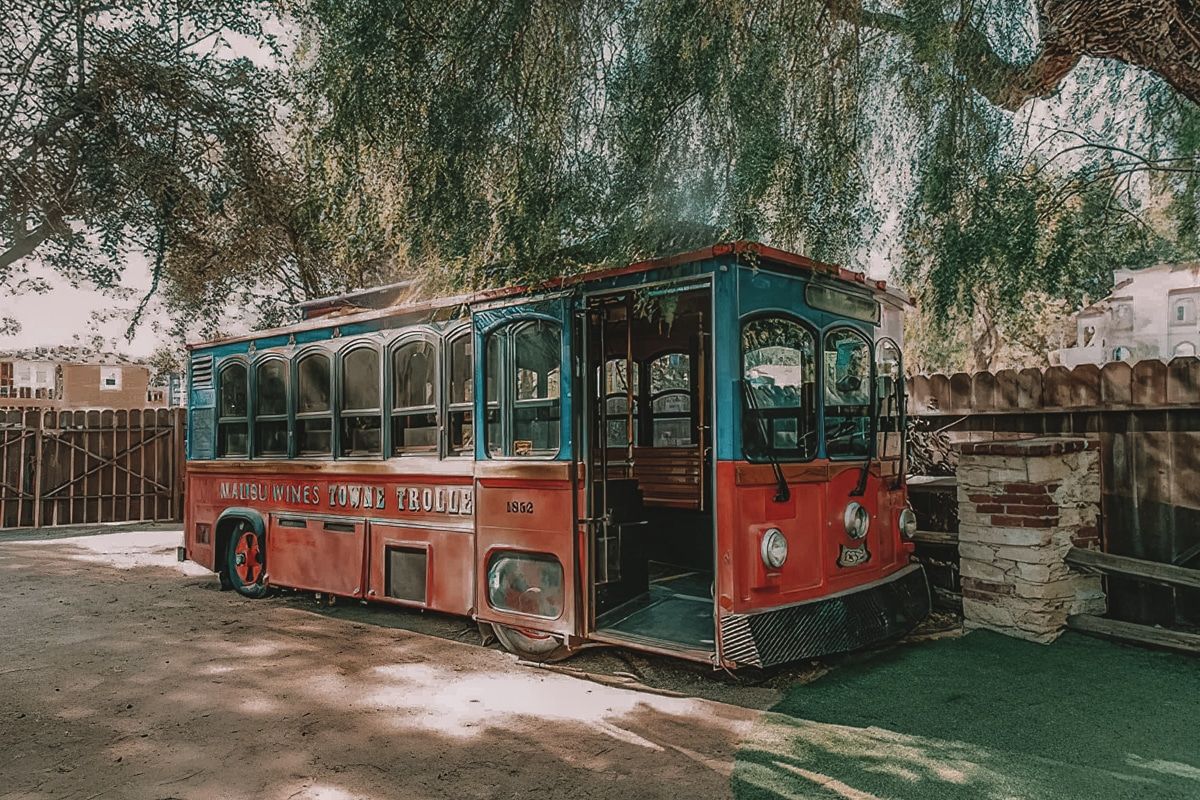 A red and blue trolley bus parked in the shade of a tree, with the words, "Malibu Wines Towne Trolley" on the side.