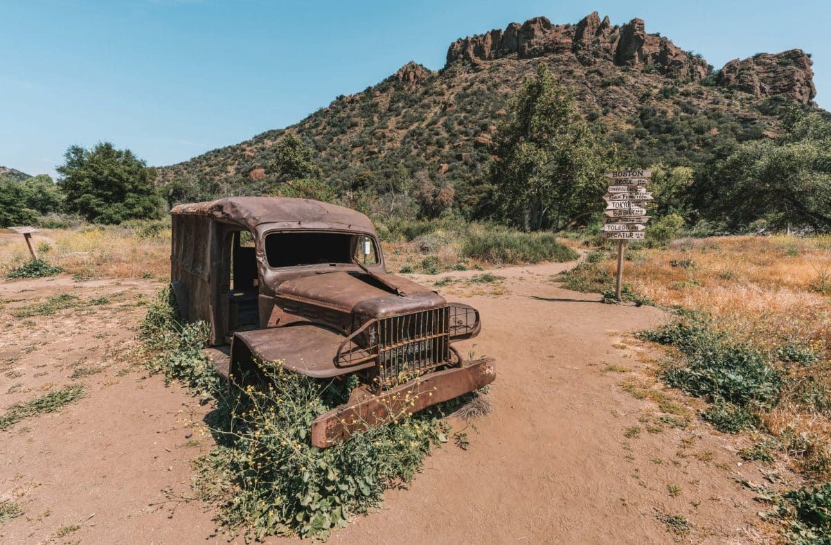 The rusted body of an antique truck, overgrown with mustard plants, and a signpost, a small, rocky mountain, and a clear blue sky in the background.