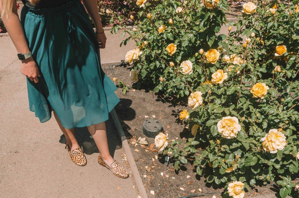 A woman wearing a turquoise dress ad cheetah-print flats seen from the waist down standing next a bush of yellow roses.
