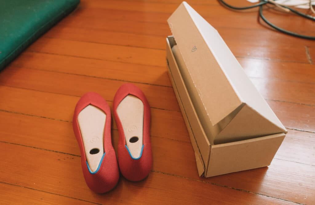 A pair of red Rothy's flats sitting on a hardwood floor with a shoebox beside them.